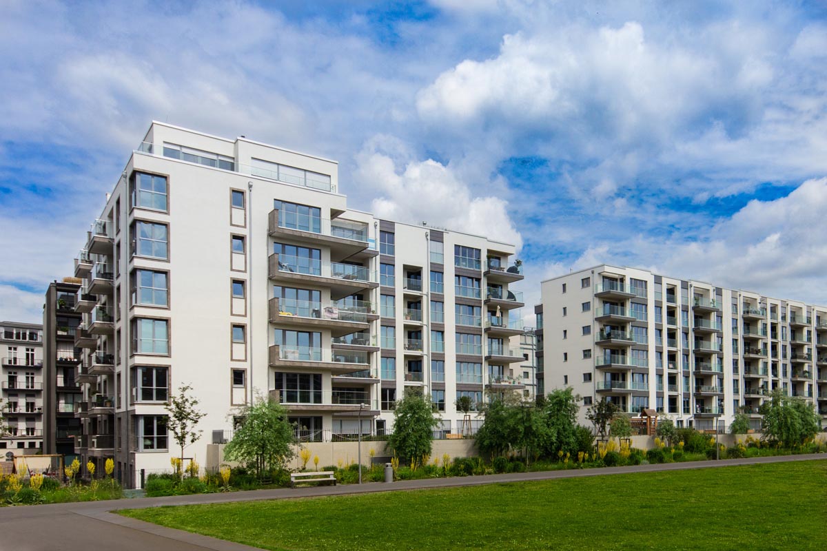 White apartment buildings with balconies, facing a green lawn under a cloudy sky.