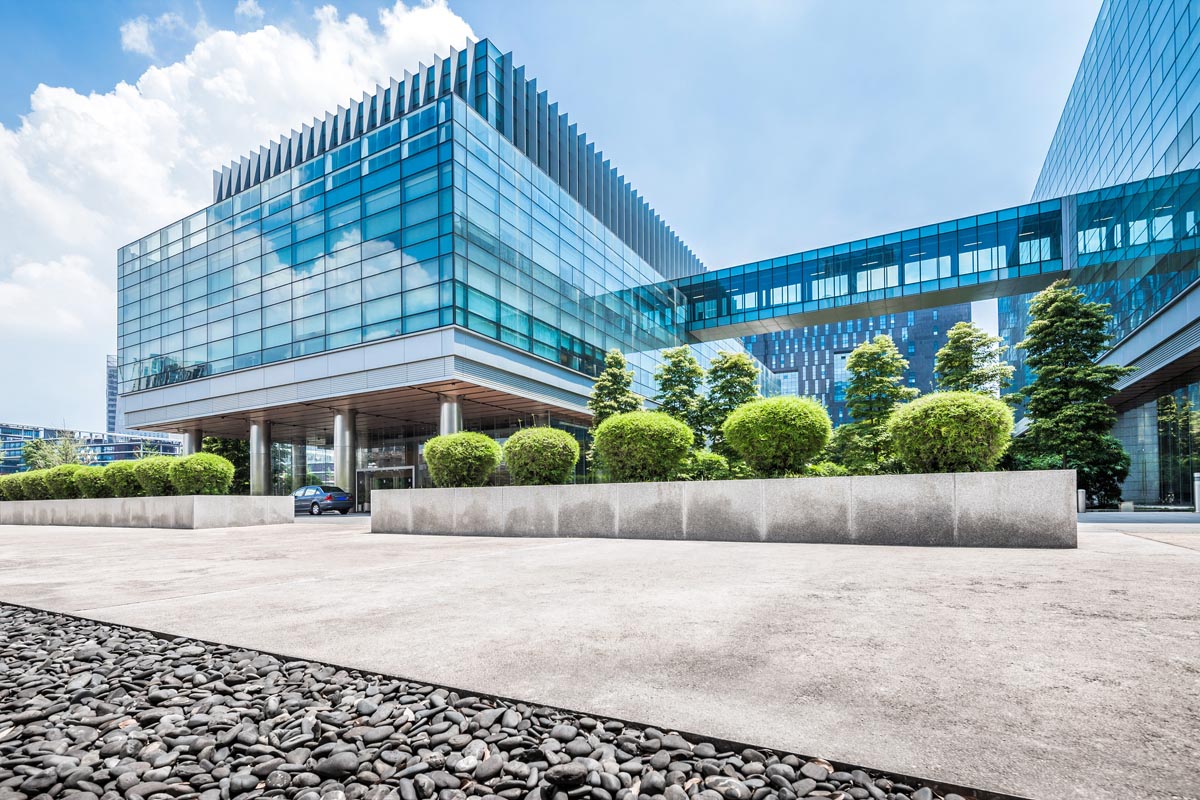 A glass office building with a skybridge, shrubs, and a wide plaza.