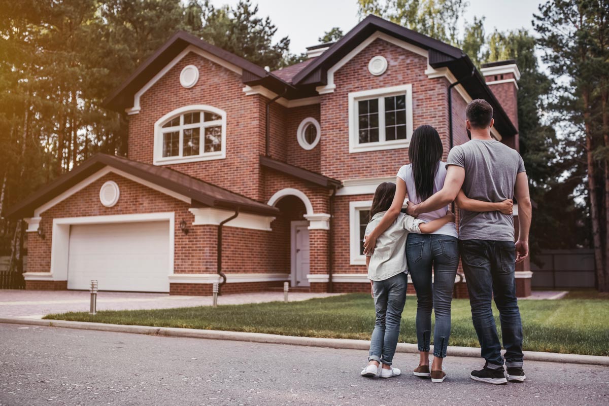 A family of three standing in the street, looking at a brick house.