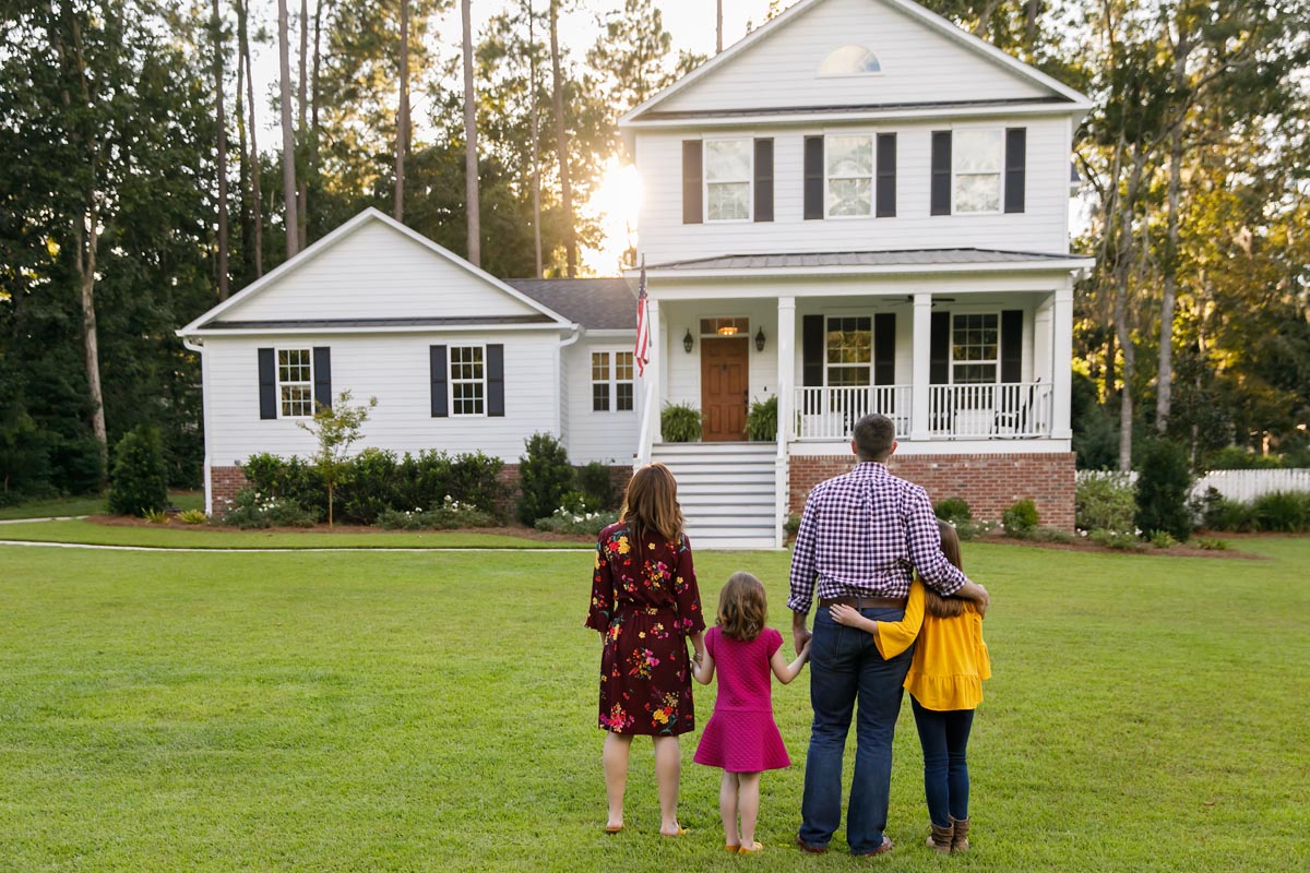 A family of four standing on a lawn, looking at a white house.
