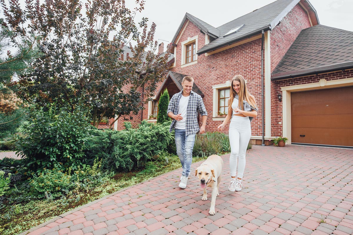 A couple walking a dog on the driveway of a brick house.