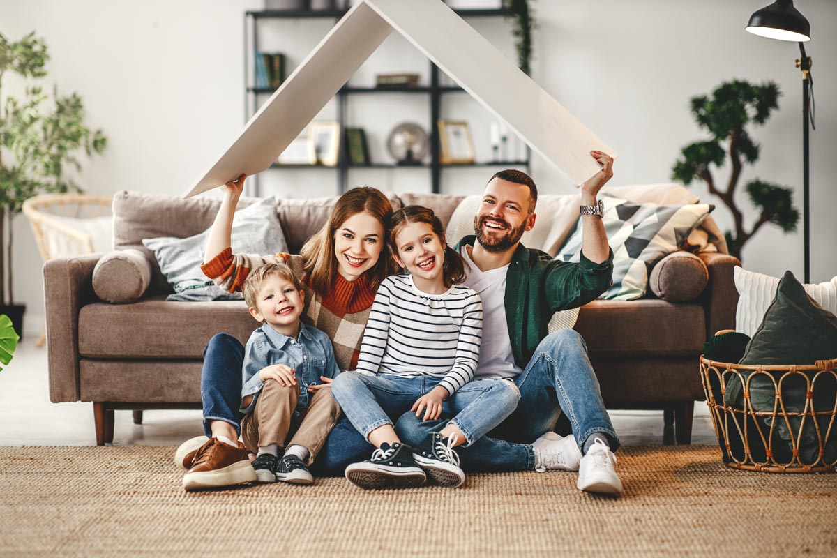 A family sitting on the floor, holding a cardboard roof over their heads.