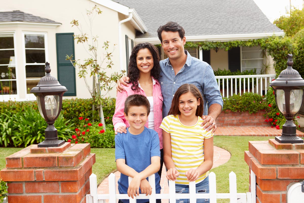A family of four standing at a white picket gate in front of a cream house.