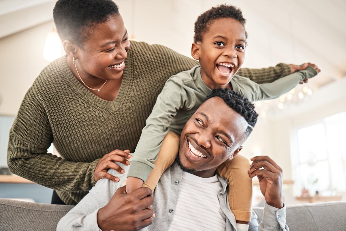A family of three smiling on a sofa, with a child sitting on a parent's shoulders.