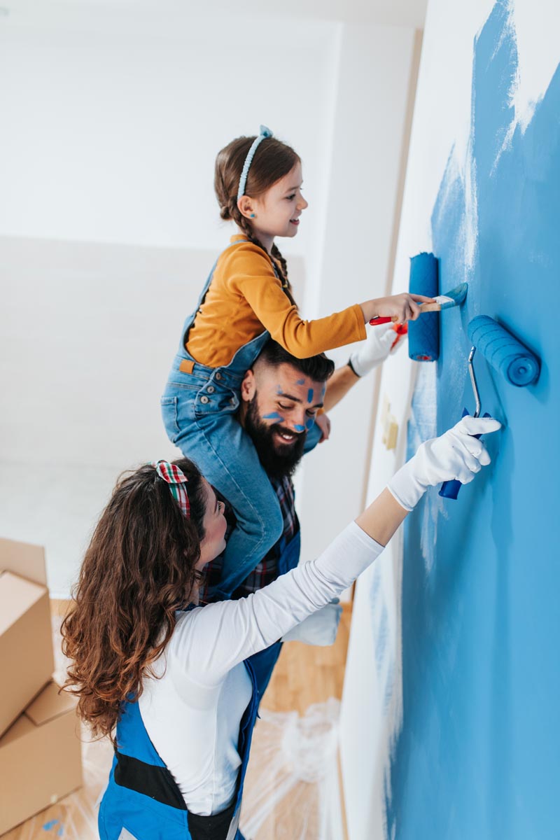 A family painting a wall blue, with a child on a parent’s shoulders.