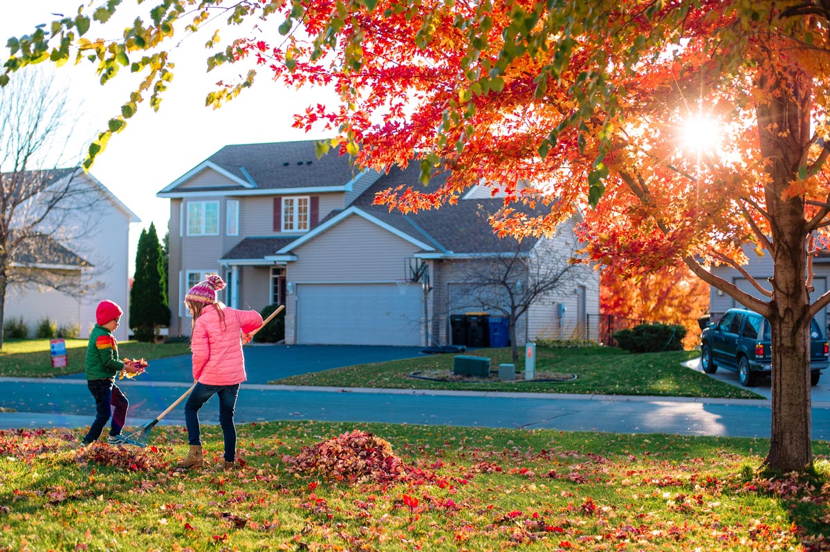 Two children raking red leaves on a lawn in front of a two-story house.