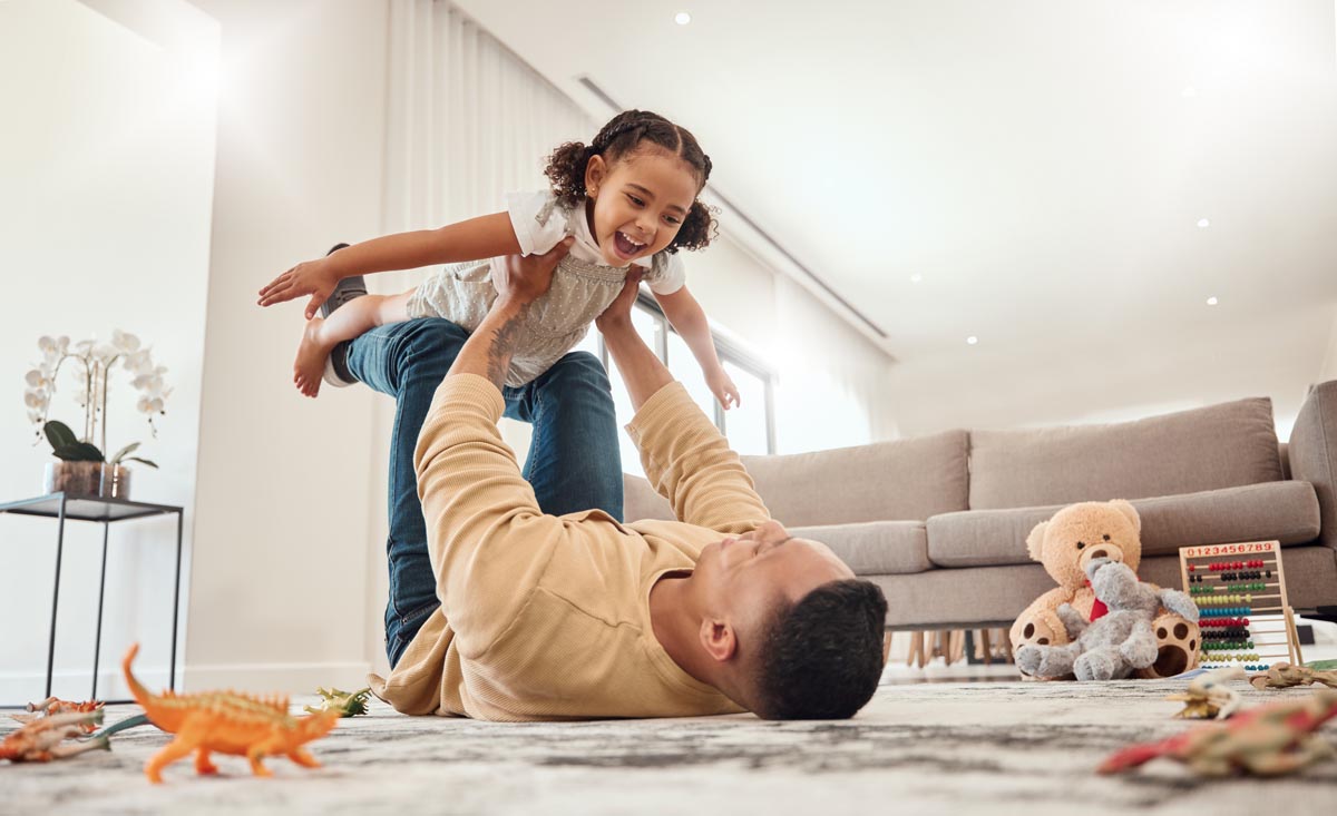 A parent lying on a rug, lifting a child into the air.