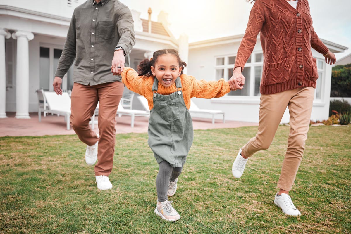 A child running across a lawn, holding hands with two adults.