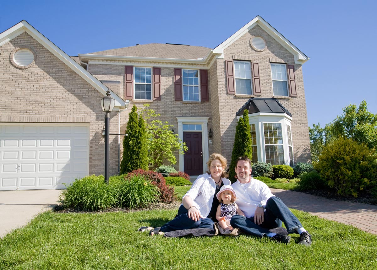 A couple and a small child sitting on the lawn in front of a brick house.