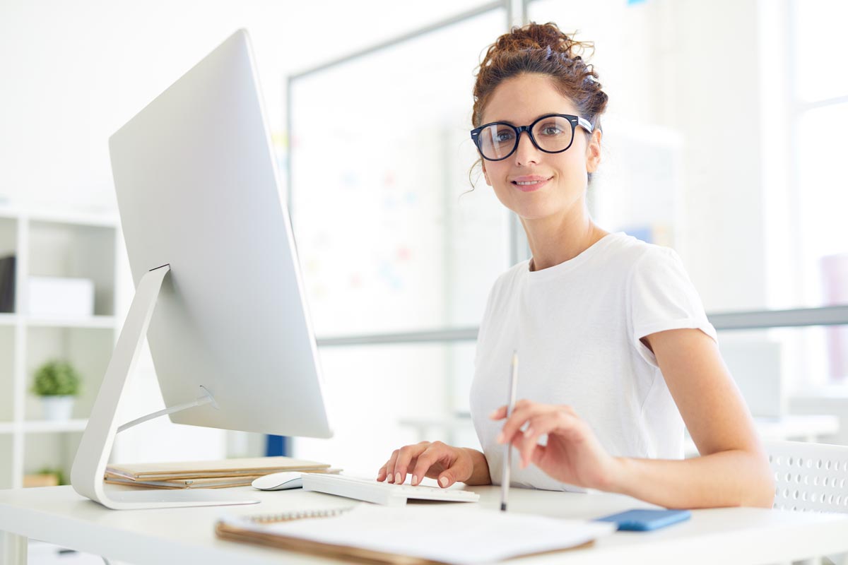 A woman at a white desk, working at a computer with a notebook in front of her.