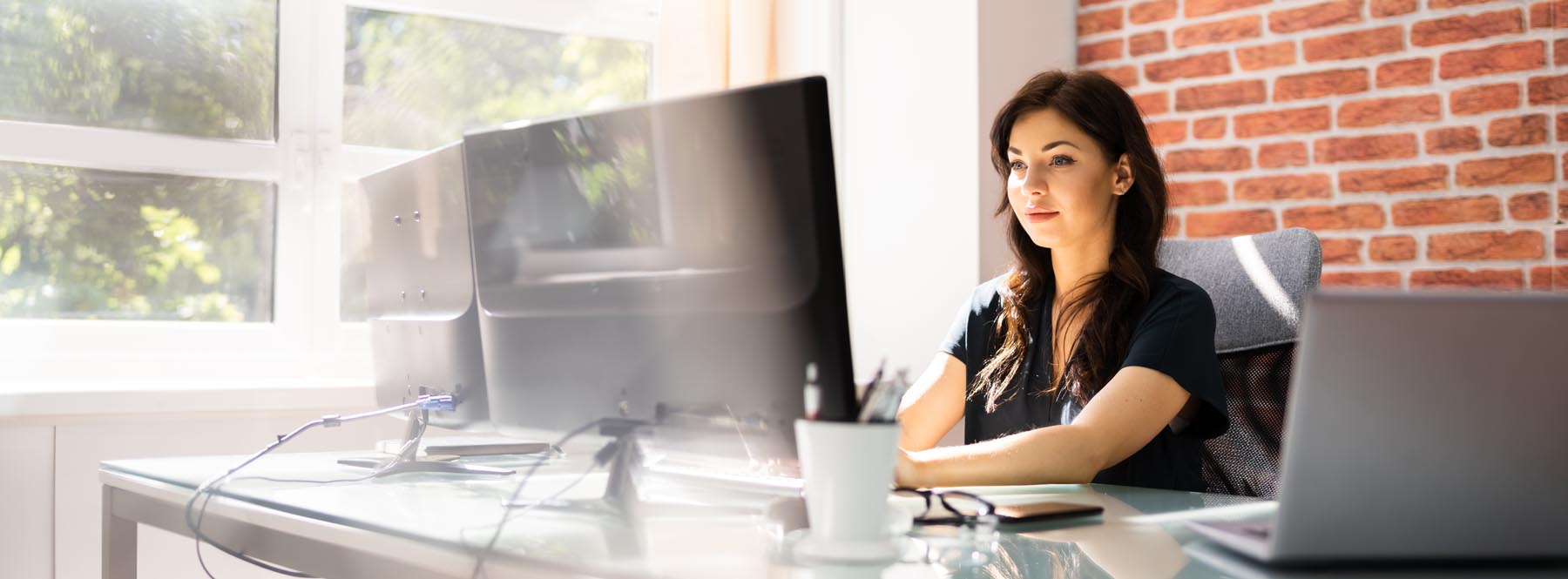 A woman working at a desk with two monitors beside a window.