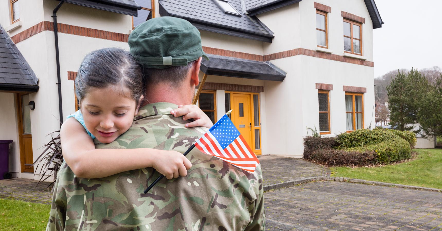 A service member in uniform hugs a child in front of a house. The child holds a small American flag.