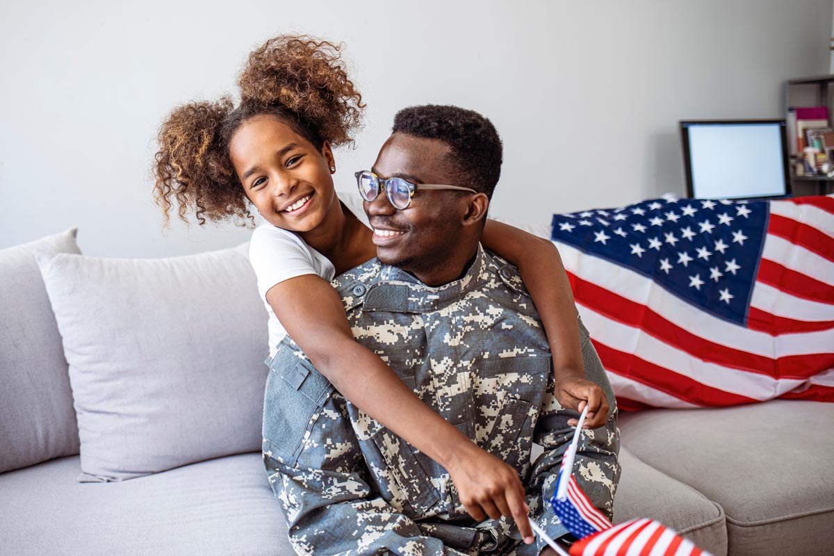 A service member in uniform sitting on a sofa with a child, and a flag draped behind them.