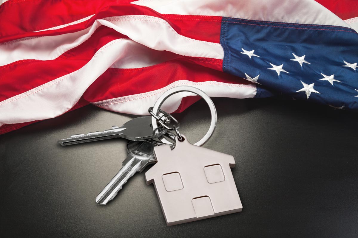 House keys and a house-shaped keychain on a table, with an American flag behind them.