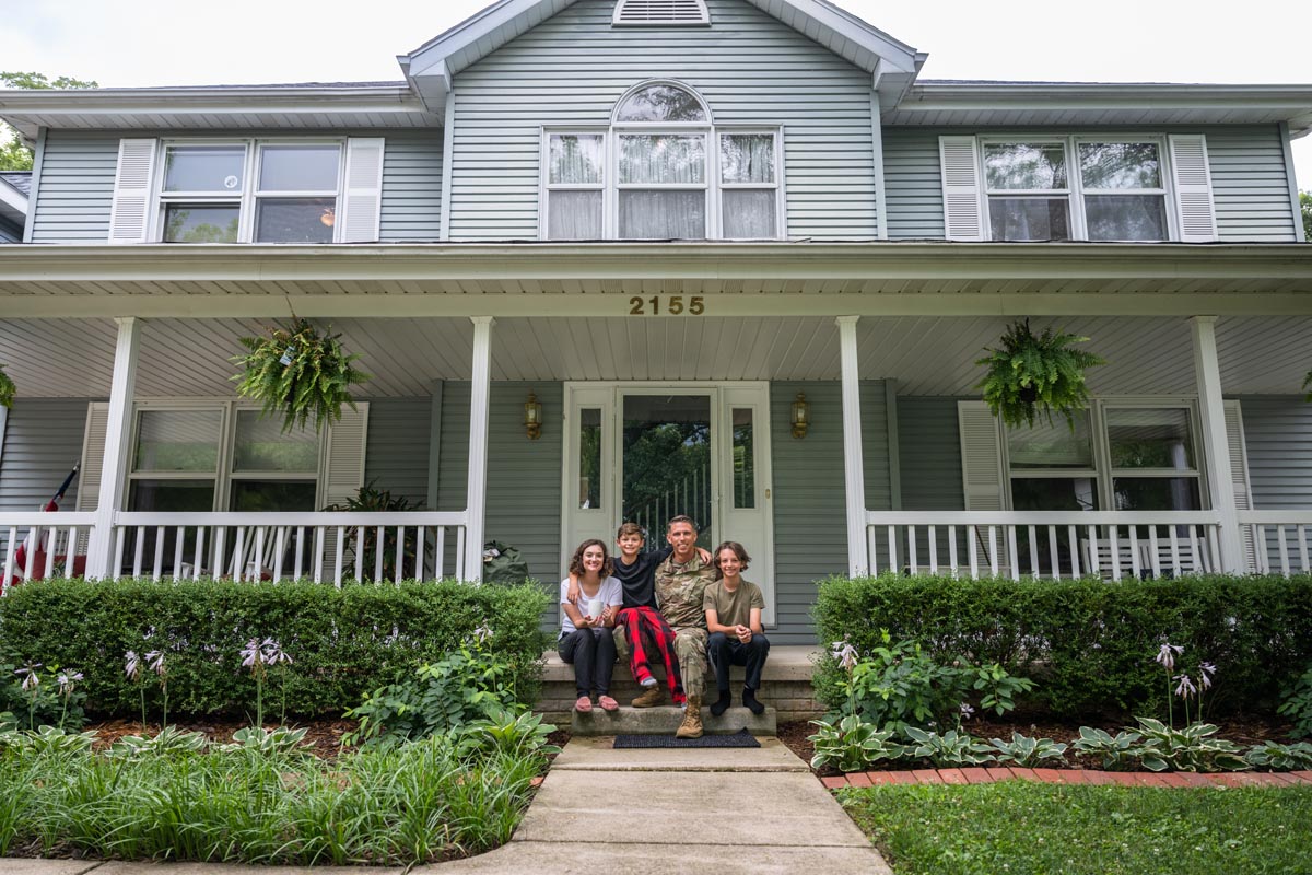 A family sitting on the front steps of a gray two-story house with a porch.