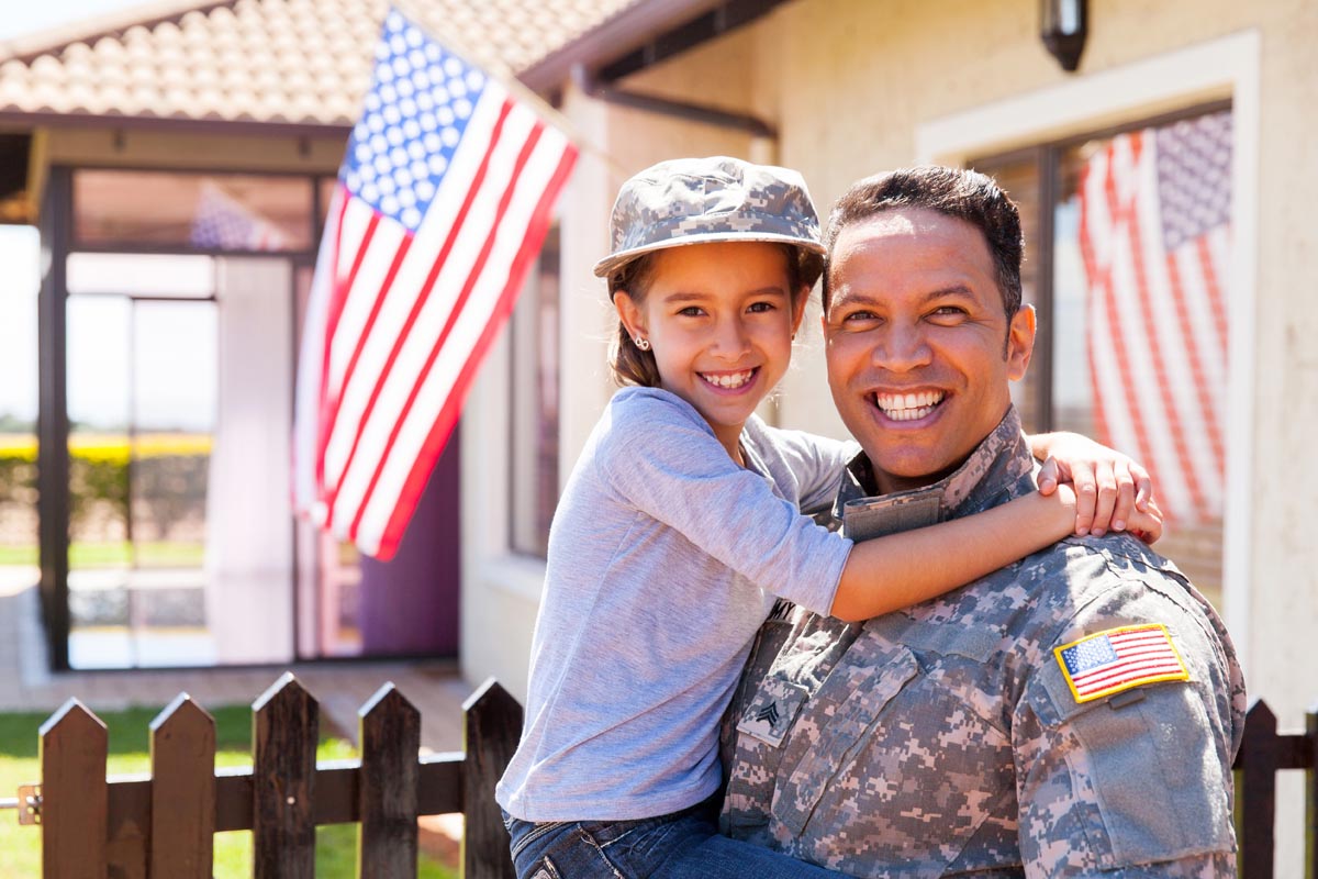 A service member holding a child in front of a house, with an American flag by a fence.