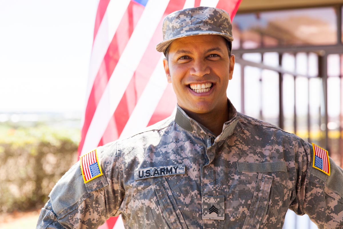 A smiling service member in a camouflage uniform and cap, standing in front of an American flag.
